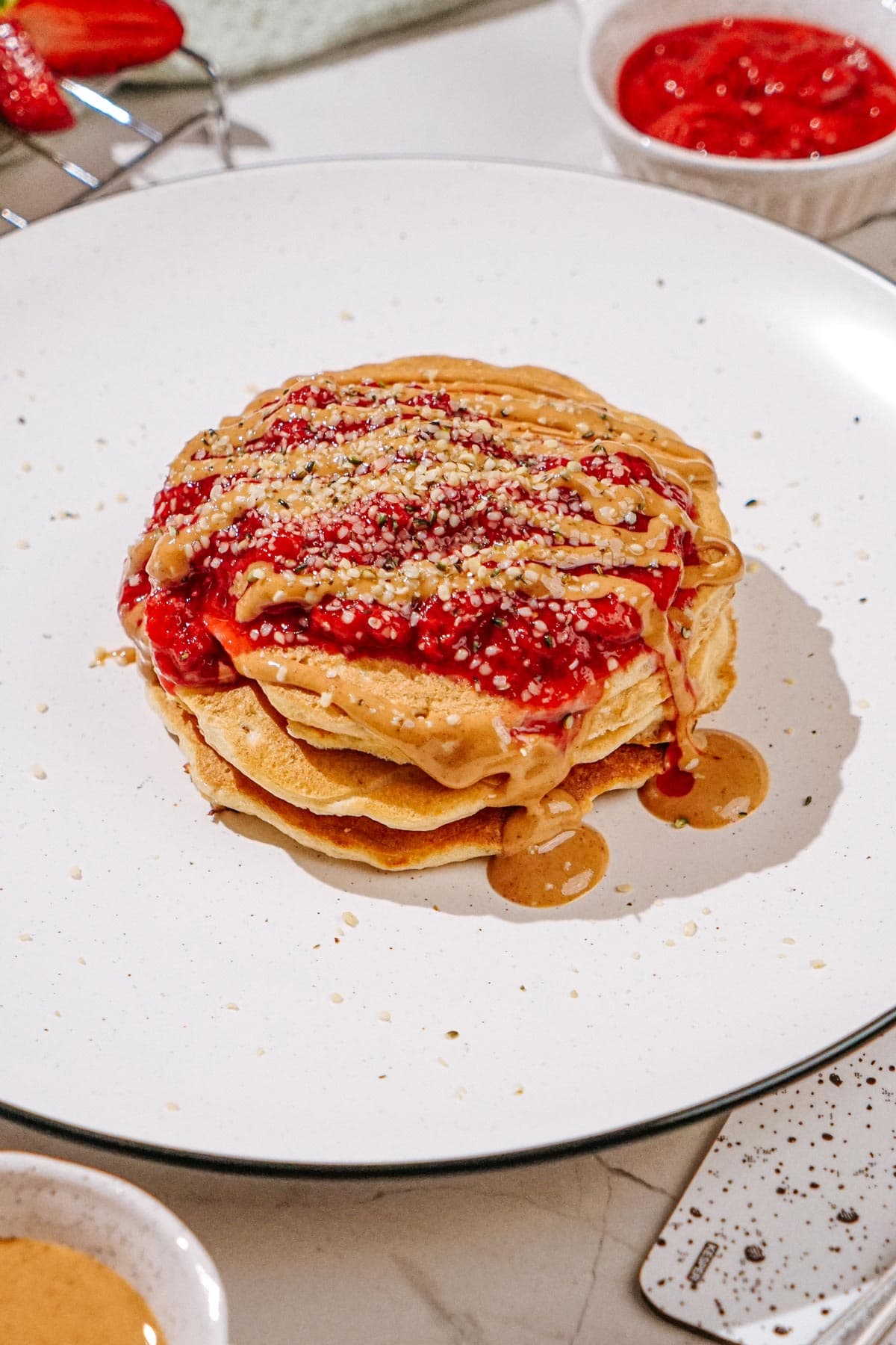 The plate with fluffy protein powder pancakes, which are drizzling nut butter, and sprinkling hemp seeds is on the light surface | Hurry The Food Up