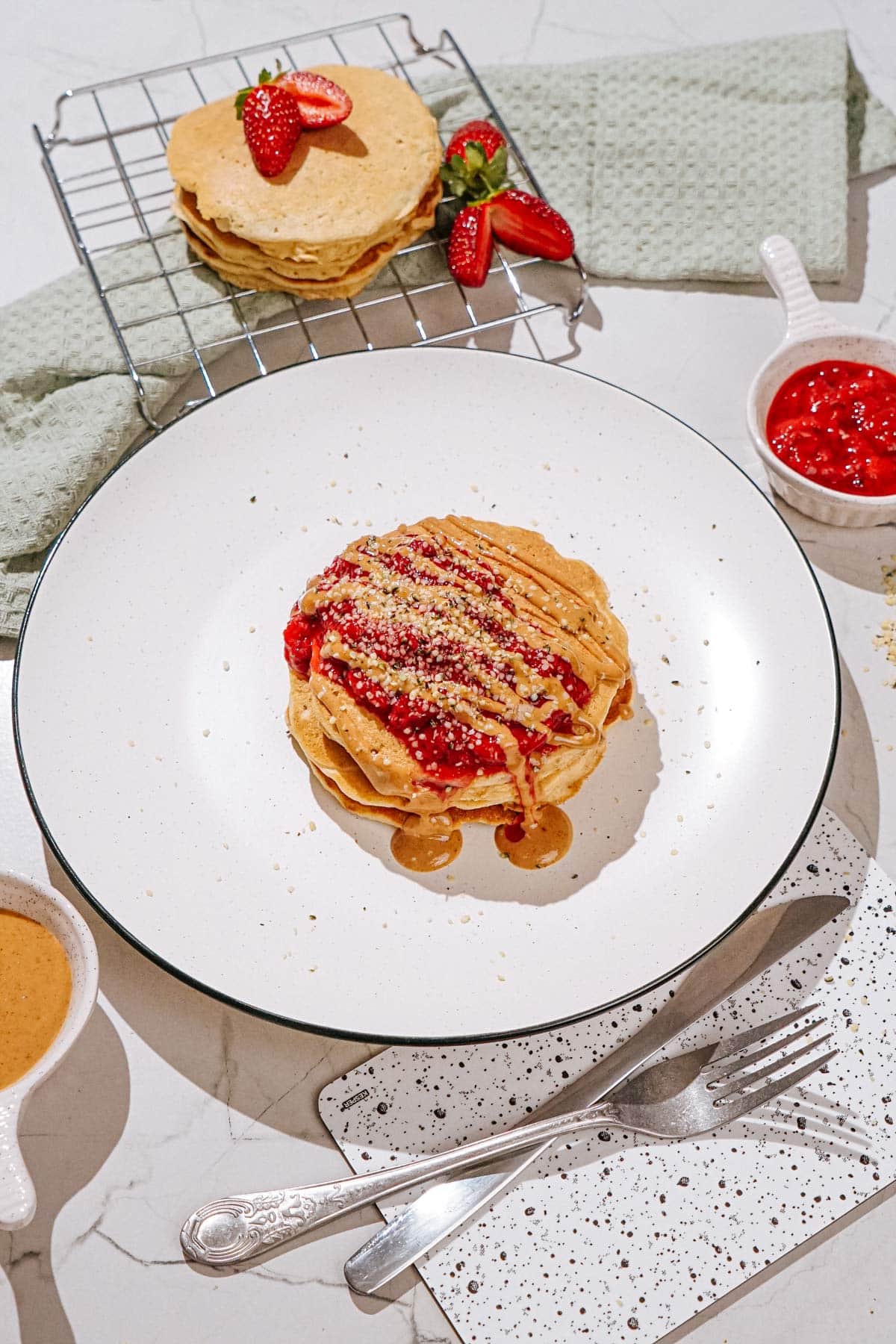 The plate with fluffy protein powder pancakes drizzled with nut butter and sprinkled with hemp seeds is on a light surface. Near it, there are some small dishes with strawberry compote, nut butter, silverware etc | Hurry The Food Up