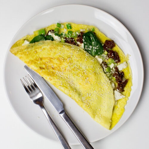 A Greek-style omelette filled with spinach, sun-dried tomatoes, feta cheese, and green onions, folded and topped with sesame seeds, served on a white plate with a fork and knife.