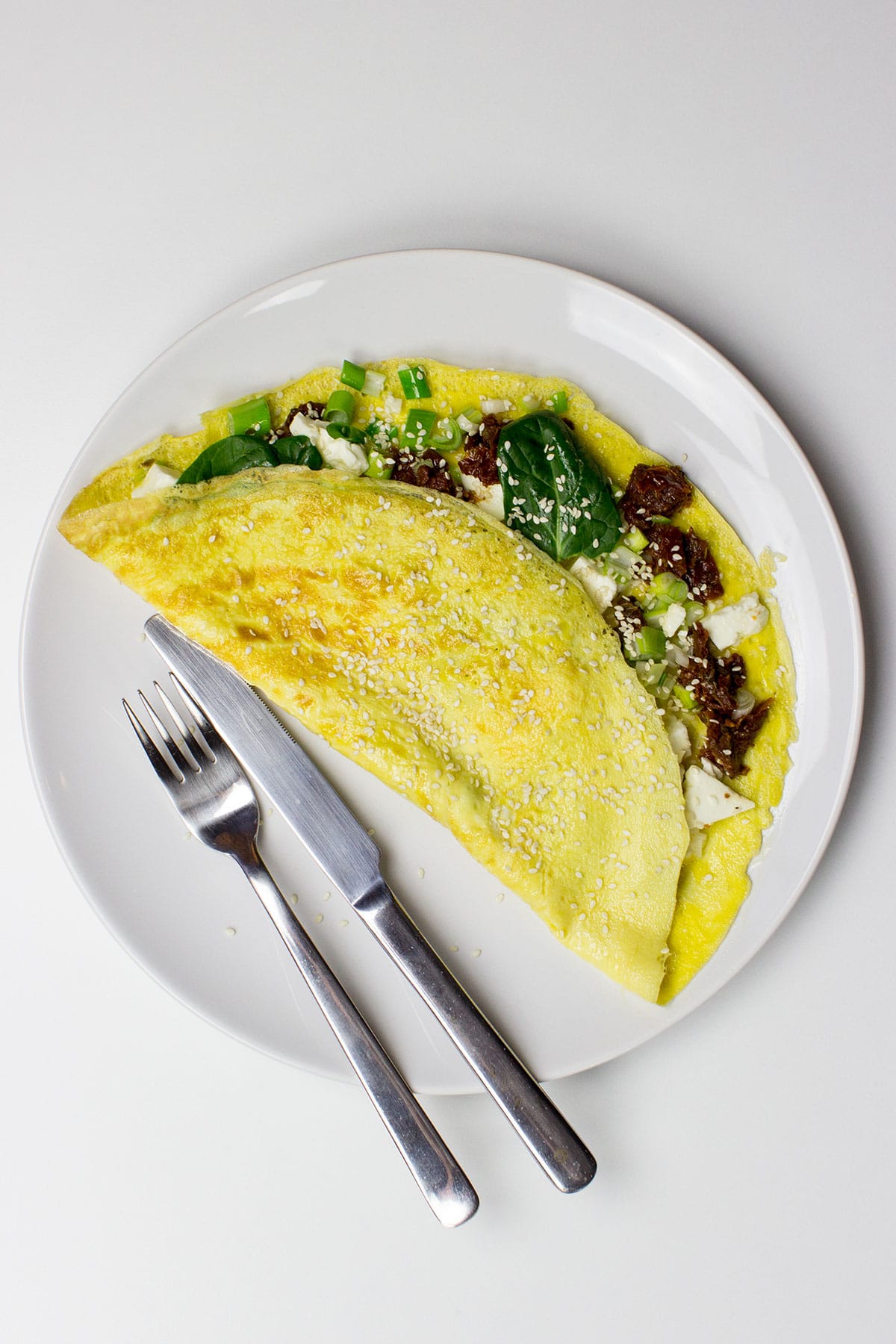 A Greek-style omelette filled with spinach, sun-dried tomatoes, feta cheese, and green onions, folded and topped with sesame seeds, served on a white plate with a fork and knife.