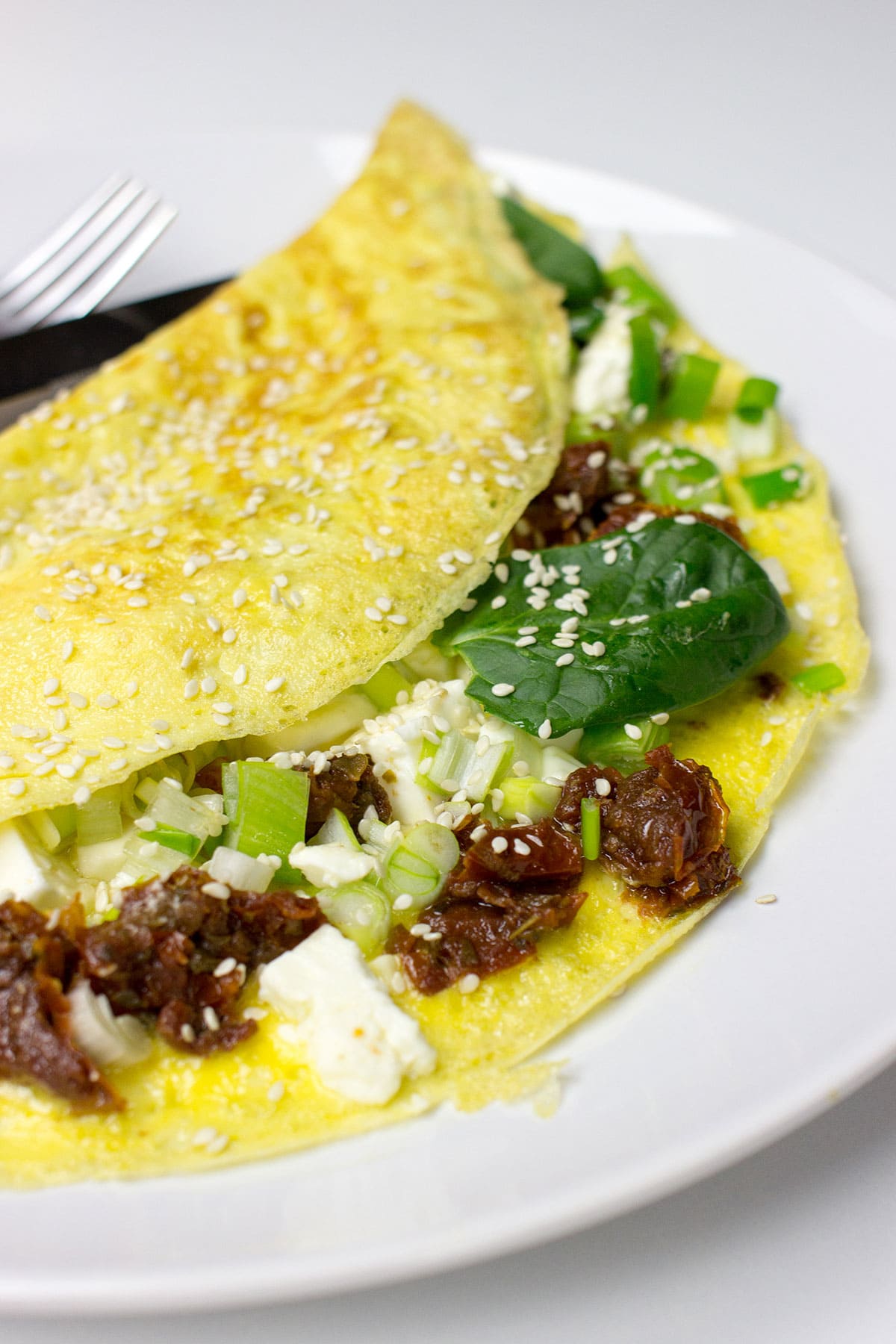 A Greek-style omelette filled with spinach, sun-dried tomatoes, feta cheese, and green onions, folded and topped with sesame seeds, served on a white plate with a fork and knife.