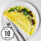 A Greek-style omelette filled with spinach, sun-dried tomatoes, feta cheese, and green onions, folded and topped with sesame seeds, served on a white plate with a fork and knife with a timestamp in the left corner.