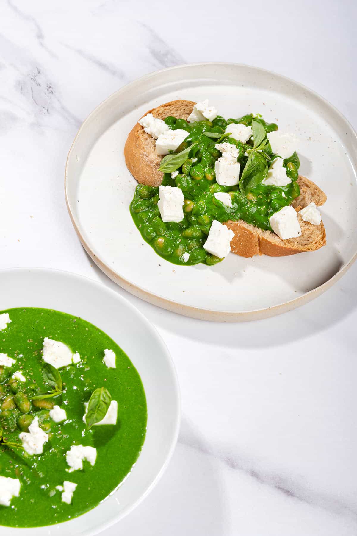Toasted bread slice that is on the white plate is topped with beans, green sauce and crumbled cheese. There is a bowl with topping along it | Hurry The Food Up