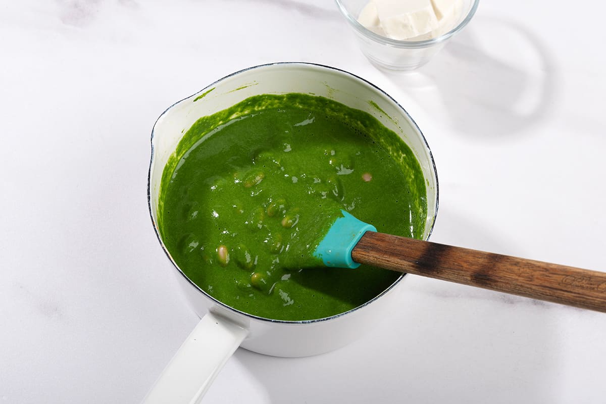 Green sauce with beans is ready in the saucepan with spatula dipped in it. There is a glass with diced feta on the side | Hurry The Food Up