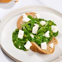 Toasted bread slice that is on the white plate is topped with beans, green sauce and crumbled cheese. There is a bread slice along it | Hurry The Food Up