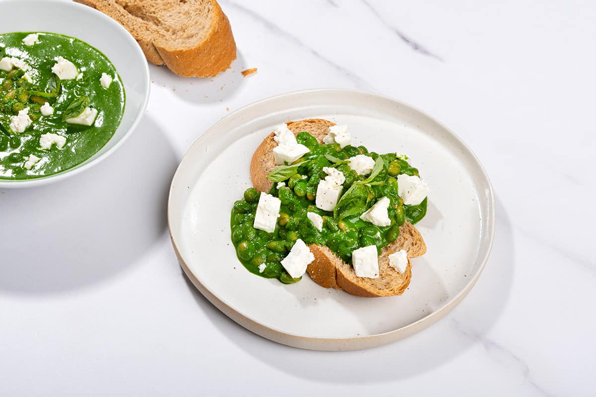 Toasted bread slice that is on the white plate is topped with beans, green sauce and crumbled cheese. There is a bowl with topping and a bread slice along it | Hurry The Food Up