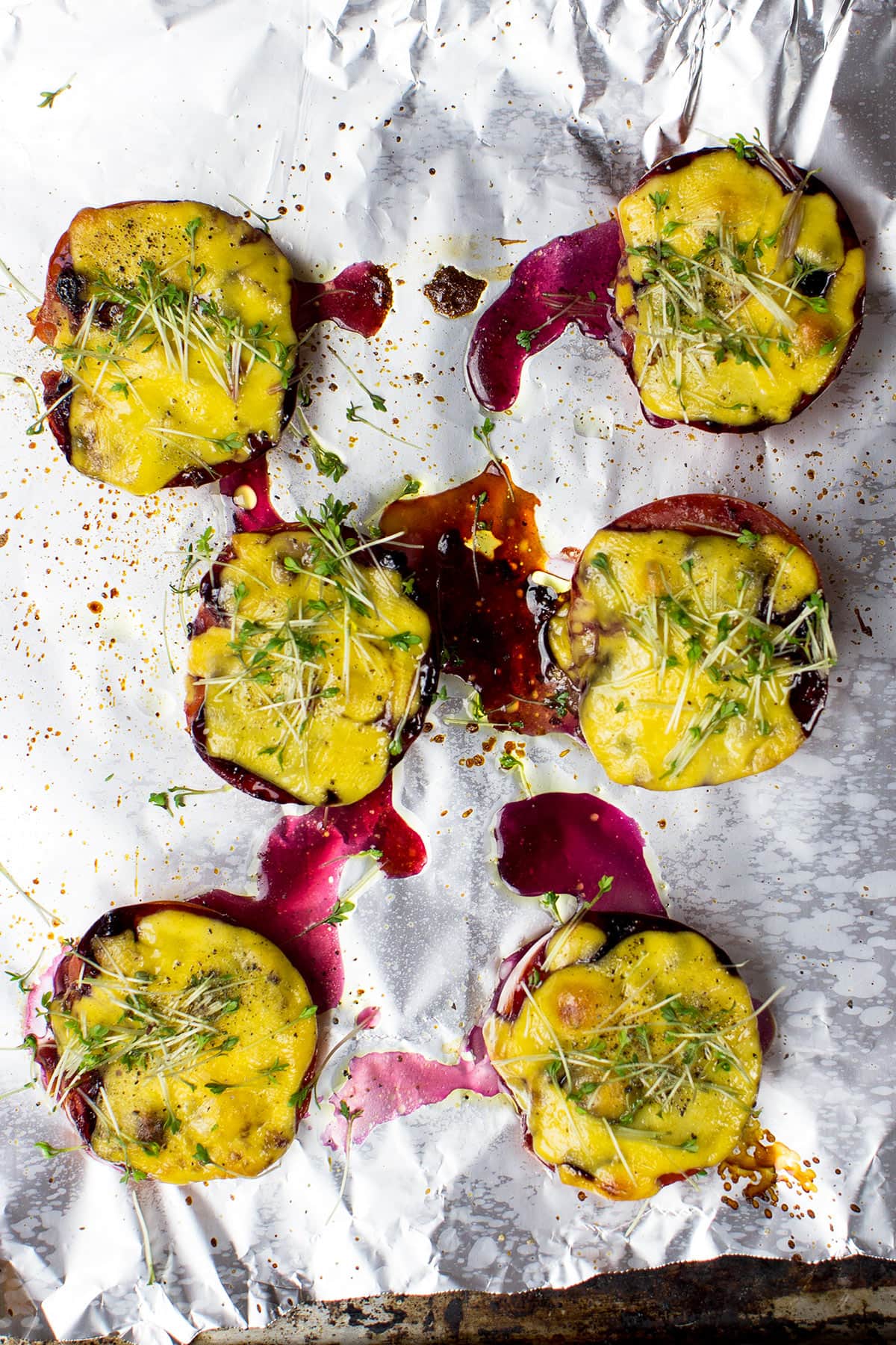 Grilled cherry tomatoes topped with melted yellow cheese and microgreens, served on a foil-lined baking tray with drizzles of savory blueberry jam.