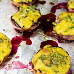 Grilled cherry tomatoes topped with melted yellow cheese and microgreens, served on a foil-lined baking tray with drizzles of savory blueberry jam.