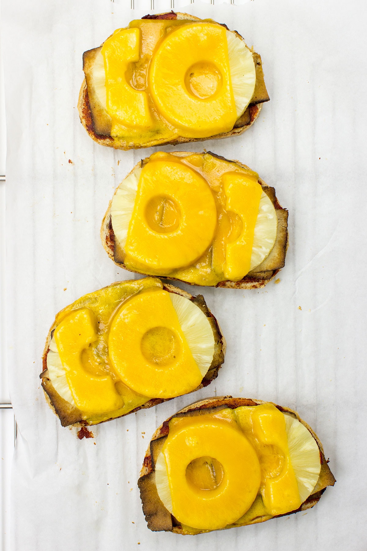 A white surface with four pieces of Hawaiian Tofu Toast topped with melted cheese and pineapple slices, arranged in a vertical line.