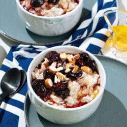 Two nice white bowls with protein oatmeal stand on the plates. Around them, there are two small spoons, glassware with honey, a plate with almonds, colorful napkin | Hurry The Food Up