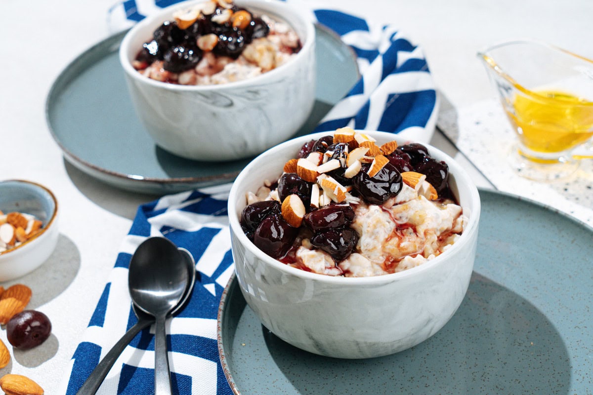 Two white bowls with protein oatmeal stand on the plates. Around them, there are small spoons, glassware with honey, a plate with almonds, colorful napkin | Hurry The Food Up