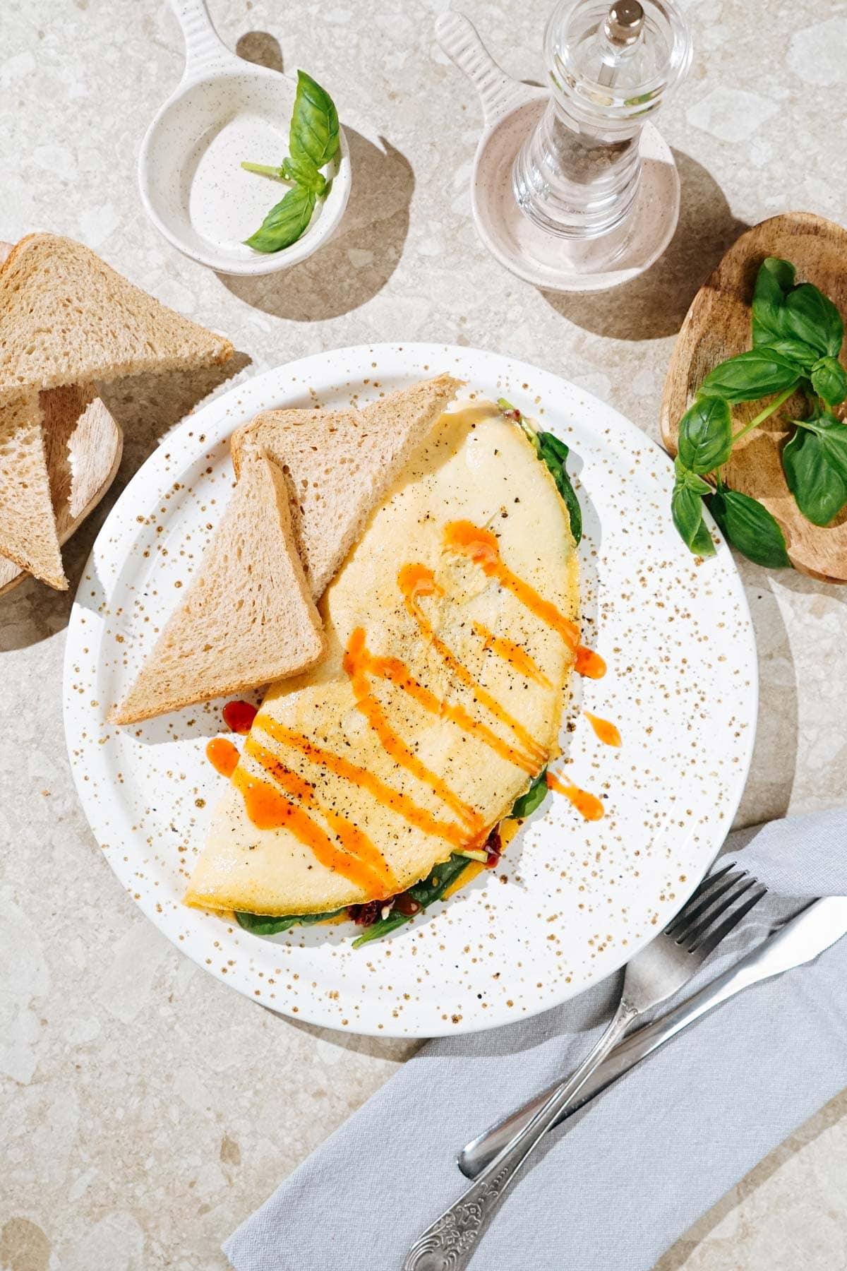 Bird's-eye view of the High Protein Omelette, topped with sriracha, salt and black pepper which is on the light plate. Two pieces of bread are on it as well. Near it there are a wooden plate with bread and basil leaves, pepper mill, fork and table napkin | Hurry The Food Up