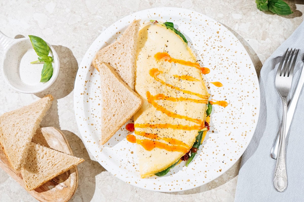 Bird's-eye view of the High Protein Omelette, topped with sriracha, salt and black pepper which is on the light plate. Two pieces of bread are on it as well. Near it there are some basil leaves, fork and table napkin | Hurry The Food Up