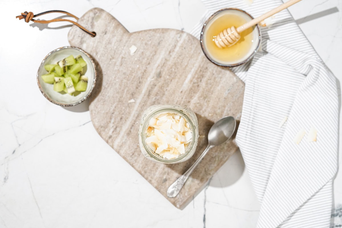 Bird's-eye view of high-protein overnight oats and spoon near it which are on the marble сut board. Near it, there are small bowls with kiwi and honey | Hurry The Food Up