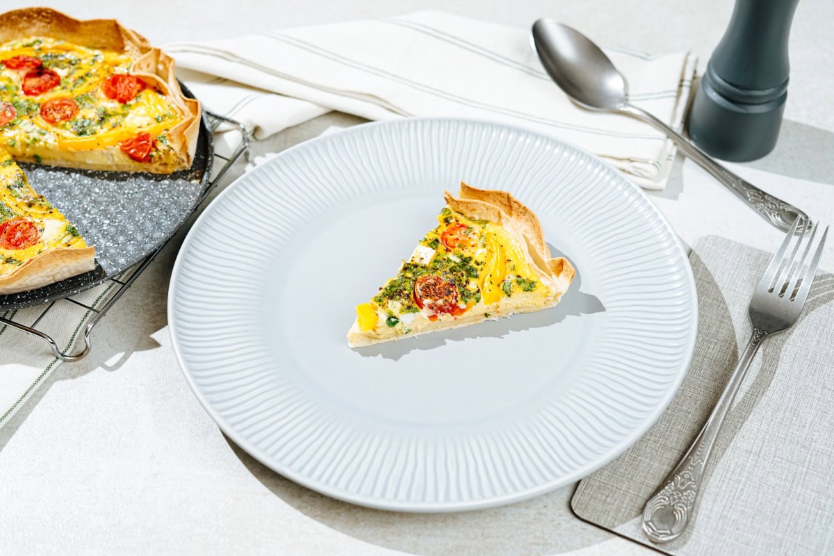 The plate with the piece of the tortilla quiche is on the white surface. There are a fork, spoon, kitchen towel, grinder, and a black plate with tortilla quiche around it | Hurry The Food Up
