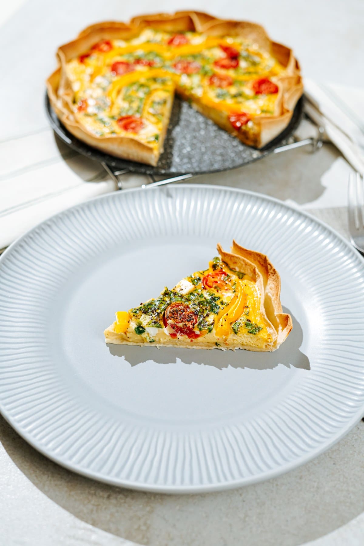 A piece of high-protein tortilla quiche lies on the white plate, and there is a fork on the right side of it. Behind the white plate, there is tortilla quiche on the black plate | Hurry The Food Up