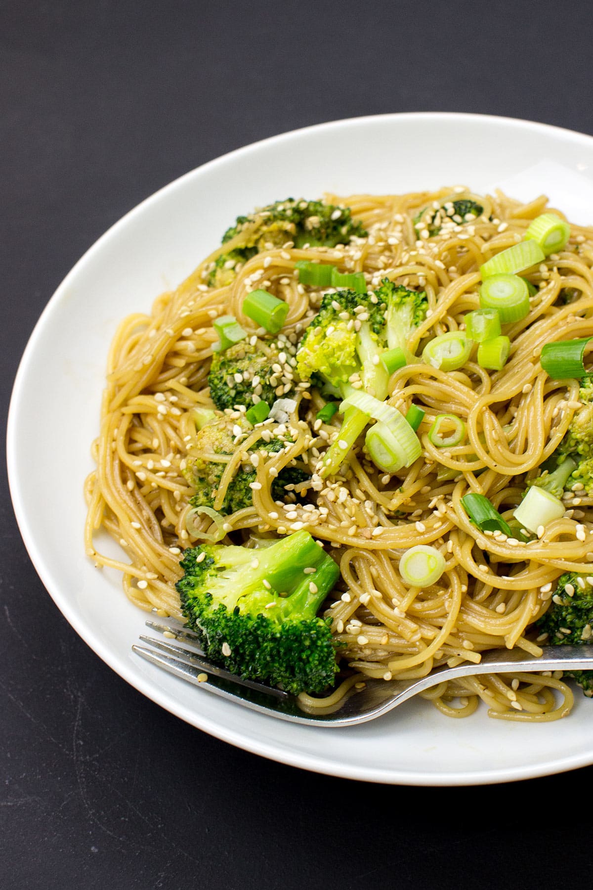 A white plate topped with Hoisin Ginger Noodles, featuring broccoli, green onions, and sesame seeds, with a fork resting on the side.