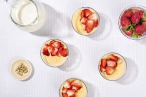 A bird’s eye view of 4 ramekins of custard dessert, a jar of milk, cardamom seeds & some strawberries | Hurry The Food Up