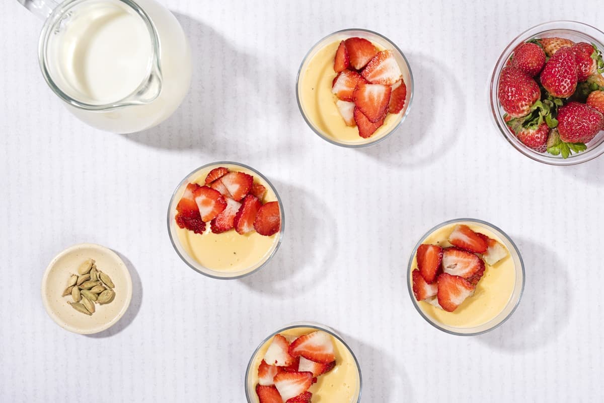 A bird’s eye view of 4 ramekins of custard dessert, a jar of milk, cardamom seeds & some strawberries | Hurry The Food Up