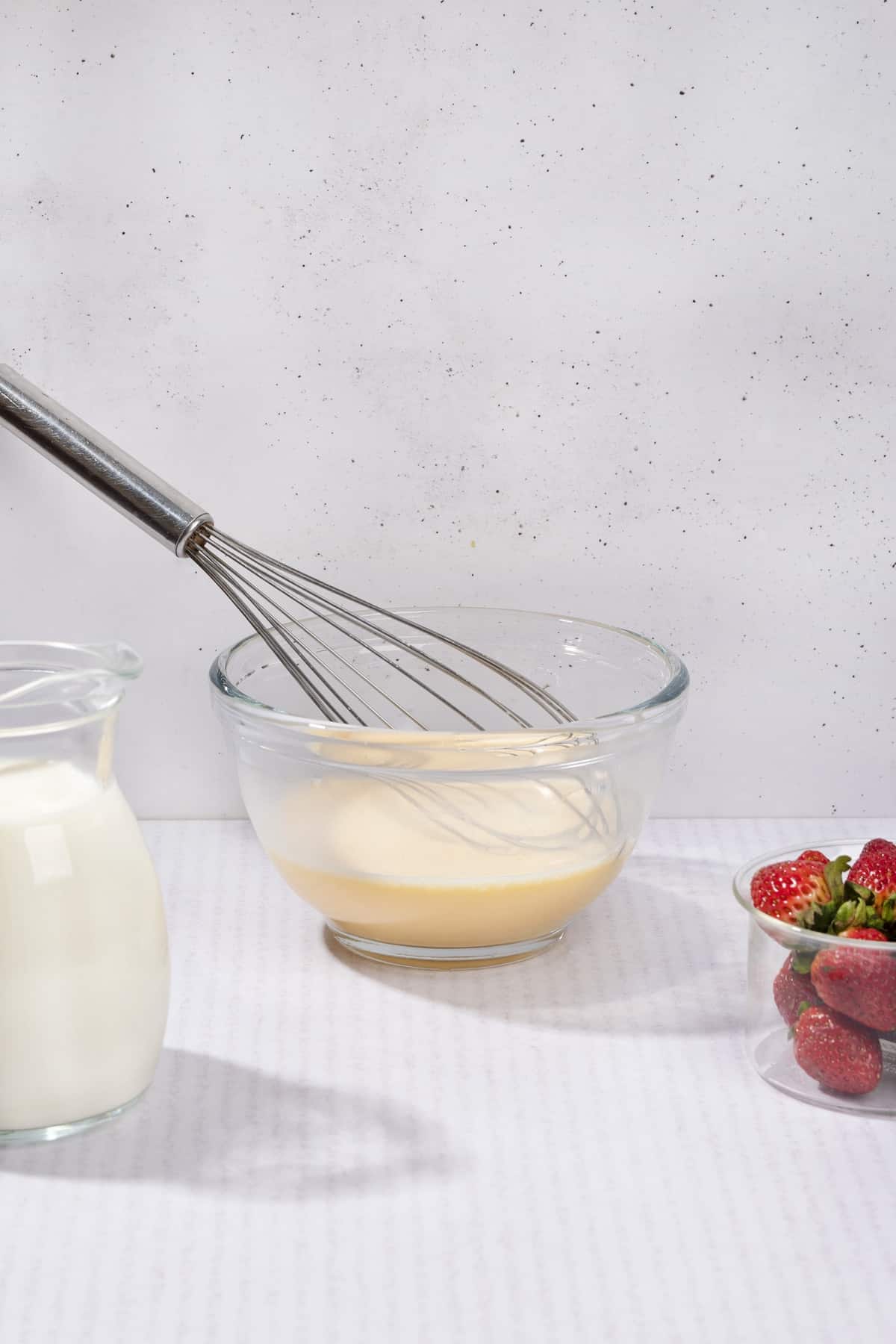 A glass bowl of uncooked custard with a whisk in it. A small bowl of strawberries and a jug of milk | Hurry The Food Up