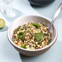 A bowl of lentil tabbouleh with a spoon on a plain surface. Visible at the top of the image is another bowl of tabbouleh , and between the two to the left is a glass of water and a slice of lime | Hurry The Food Up