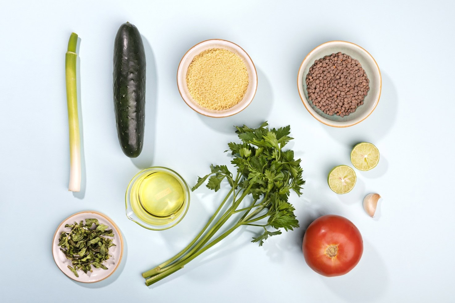 The ingredients for a tabbouleh are laid out, including a green onion, cucumber, couscous, green lentils, chopped mint, olive oil, parsley, a tomato, a lime sliced in two halves and a clove of garlic | Hurry The Food Up