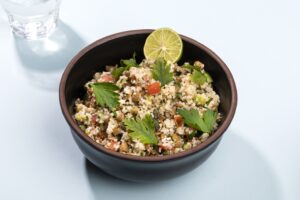A bowl of lentil tabbouleh garnished with parsley and lime, on a white surface with a glass of water in the left hand corner | Hurry The Food Up