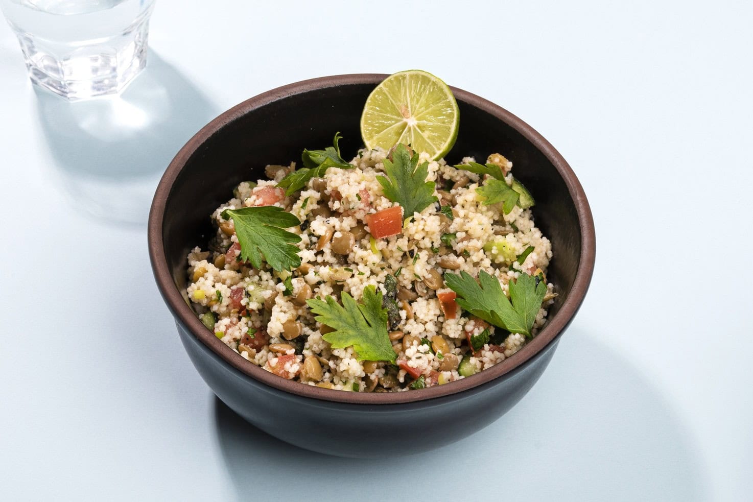 A bowl of lentil tabbouleh garnished with parsley and lime, on a white surface with a glass of water in the left hand corner | Hurry The Food Up