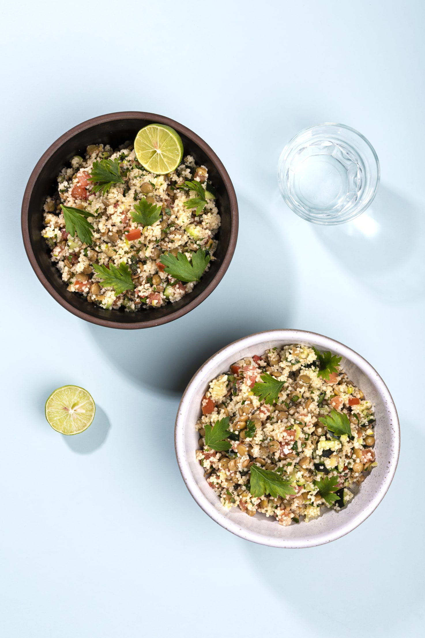 Two bowls of lentil tabbouleh from birds eye view, one in a brown bowl one in a white bowl, alongside a glass of water and half a lime | Hurry The Food Up