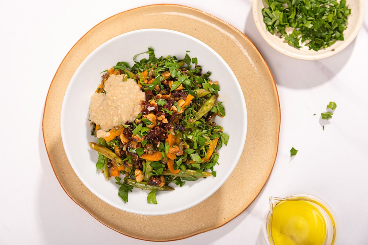 Bird's-eye view of lentils with vegetables that are served in the white plate that stands on the beige plate which is on the table. Next to it there is a plate with chopped parsley and olive oil | Hurry The Food Up