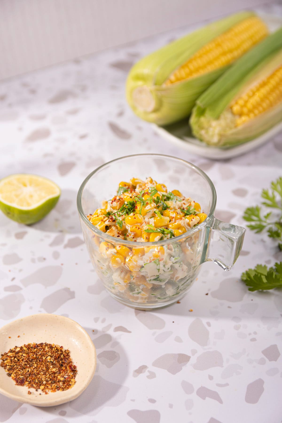 Mexican corn salad is in the glass cup on the table. Next to it there are a white plate with two corn cobs, lime half, cilantro leaves, and a plate with red pepper flakes | Hurry The Food Up