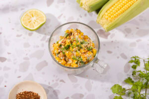 Mexican corn salad is in the glass cup on the table. Next to it there are a white plate with two corn cobs, lime half, cilantro leaves, and a plate with red pepper flakes | Hurry The Food Up