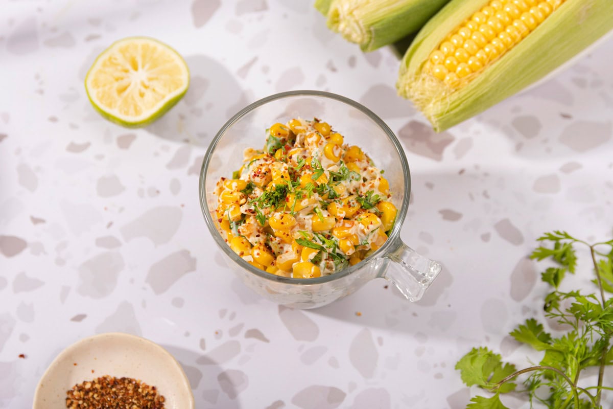 Mexican corn salad is in the glass cup on the table. Next to it there are  two corn cobs, lime half, cilantro leaves, and a small plate with red pepper flakes | Hurry The Food Up