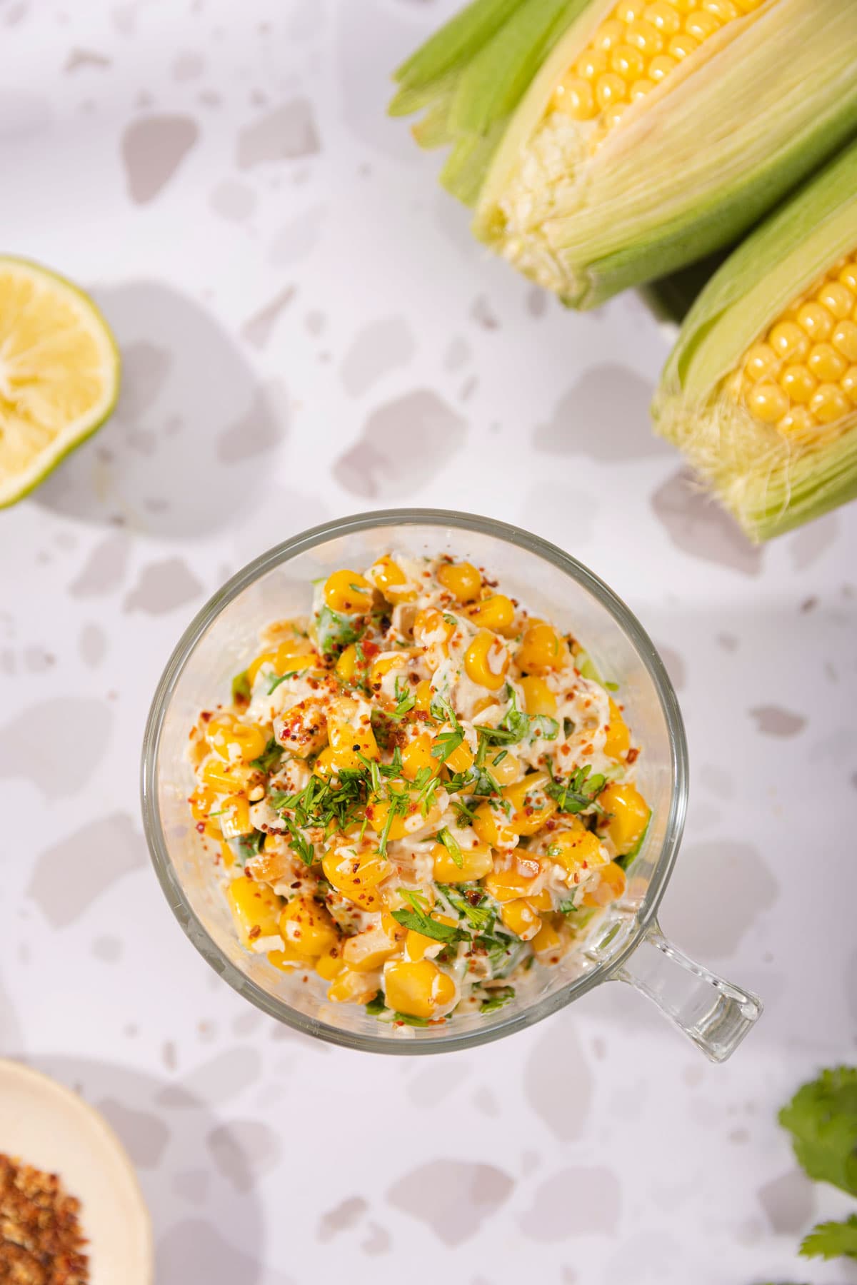 Birdeye view of the Mexican corn salad that is in the glass cup on the table. Next to it there are corn cobs, lime half, cilantro leaves, and a plate with red pepper flakes | Hurry The Food Up