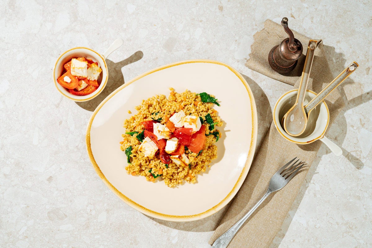 Easy high-protein millet pilaf topped with baked feta is on the light plate. There is a dish with baked vegetables and feta, two spoons, fork next to the plate | Hurry The Food Up