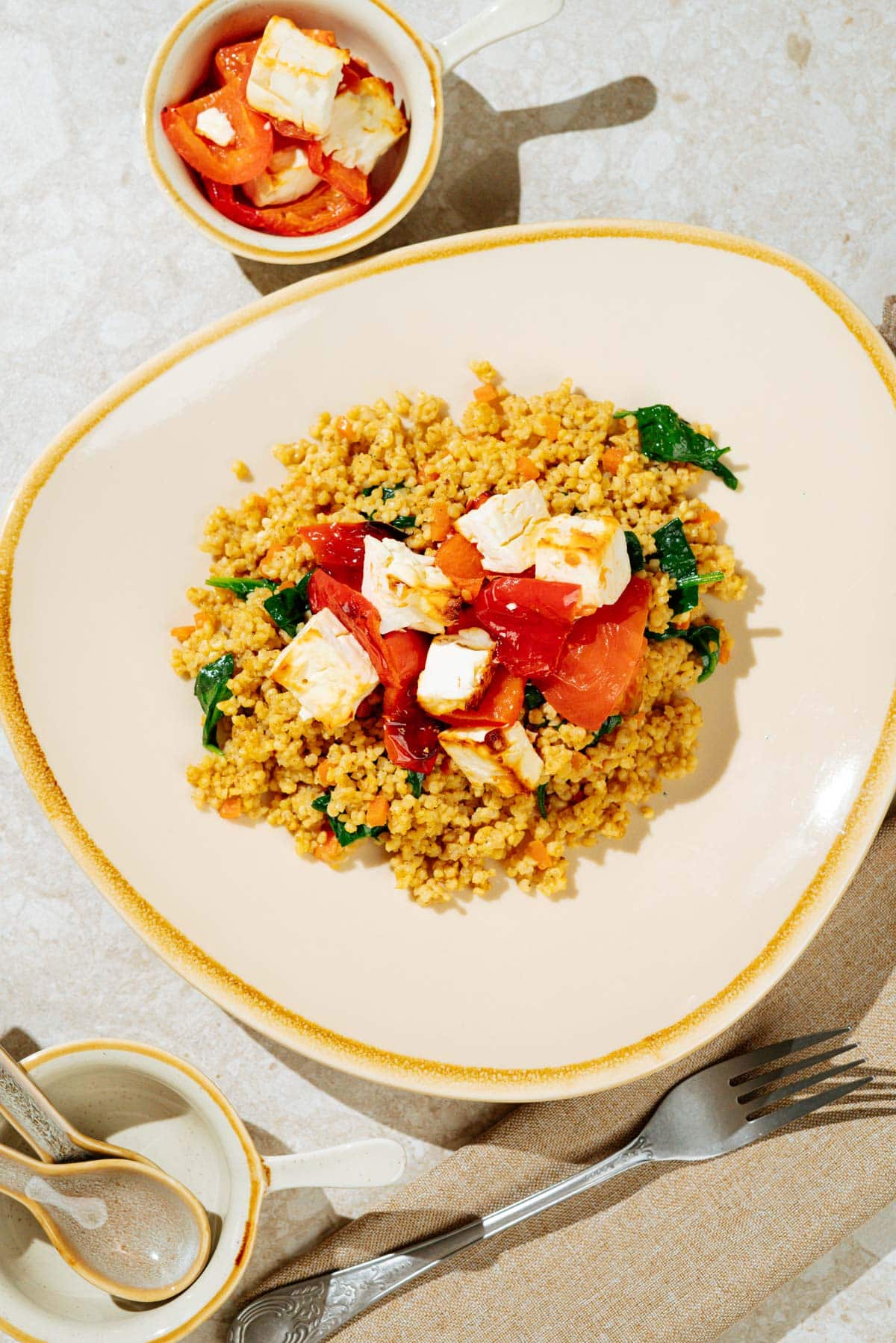 Easy high-protein millet pilaf topped with baked feta is on the light plate. There is a dish with baked vegetables and feta, two spoons, fork next to the plate | Hurry The Food Up