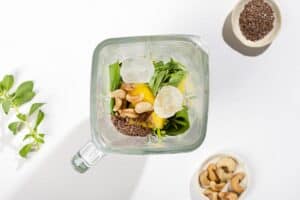 A blender filled with ingredients for a smoothie and ice cubes. Flax seeds and cashews next to it | Hurry The Food Up