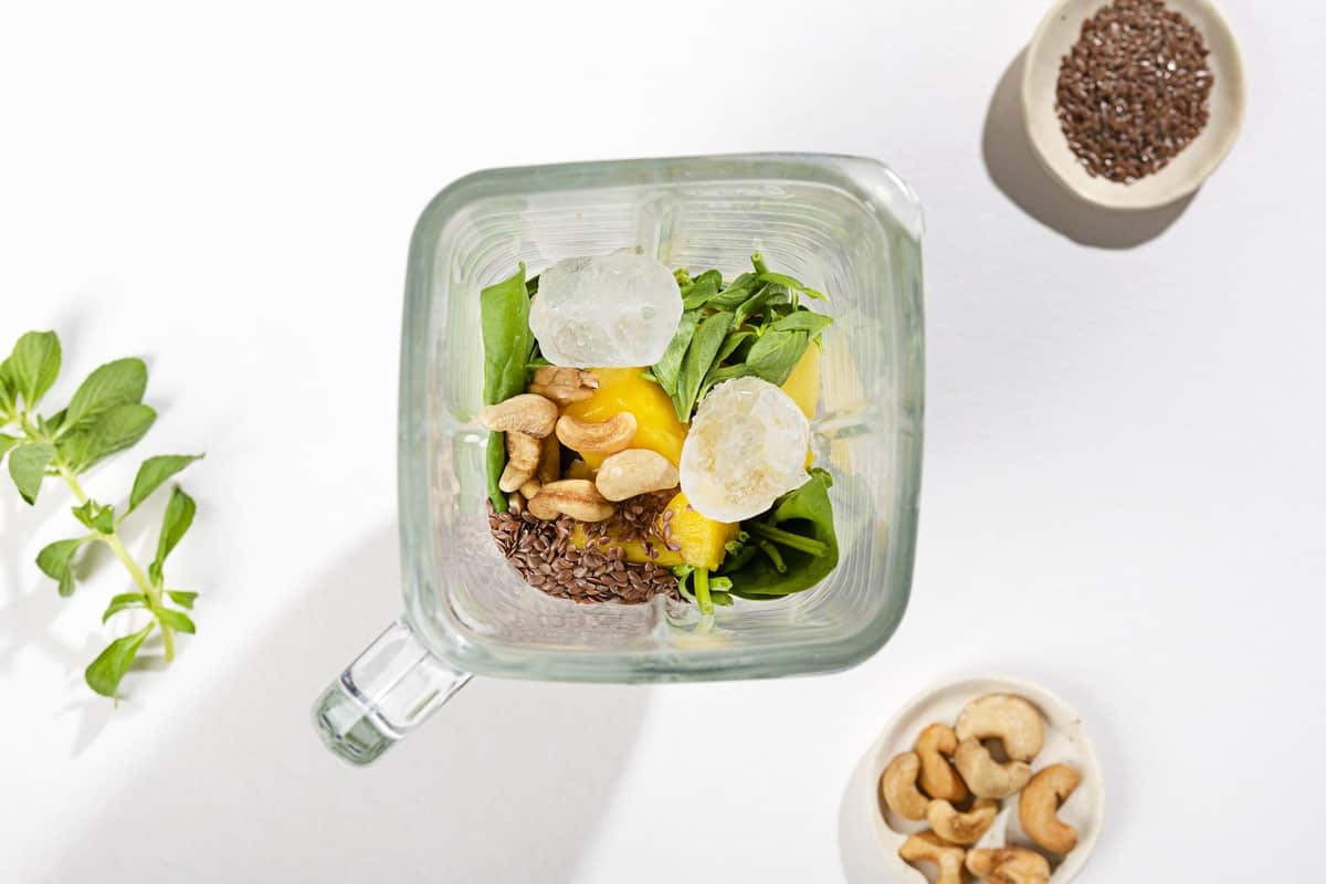 A blender filled with ingredients for a smoothie and ice cubes. Flax seeds and cashews next to it | Hurry The Food Up