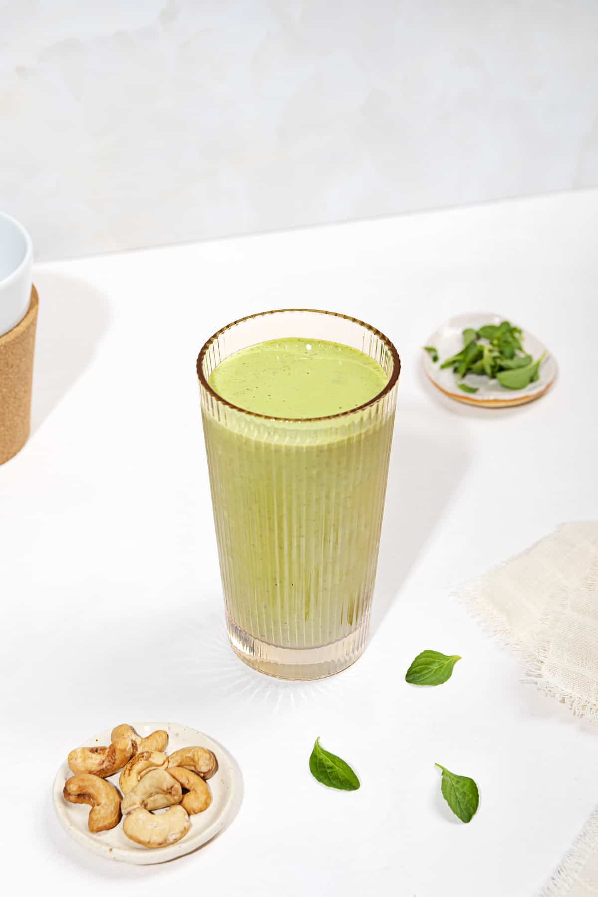 A cup filled with green smoothie on a white surface. Beside it, two small trays with cashews and mint| Hurry The Food Up