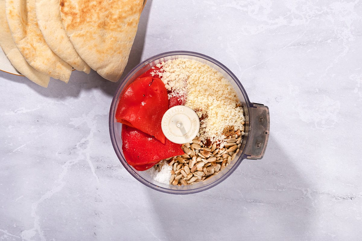 The ingredients like roasted bell peppers, roasted sunflower seeds, panko, cumin, lemon juice, sugar, paprika, salt and chili flakes are placed into the food processor. There is a plate with flatbreads on the side | Hurry The Food Up