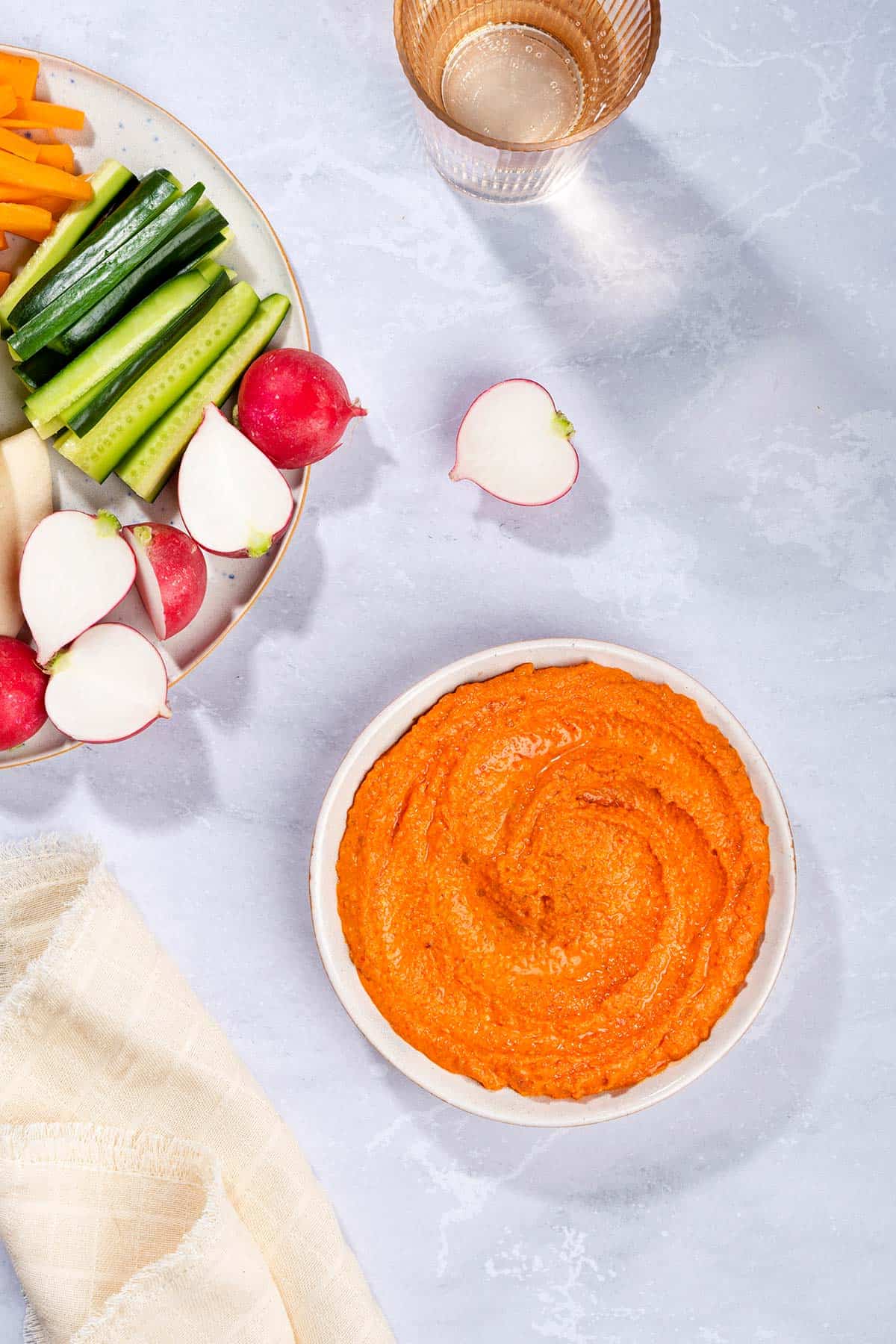 A birds eye view of a bowl with muhammara dip, a plate with vegetables and flatbreads, a glass of water and a tea towel | Hurry The Food Up