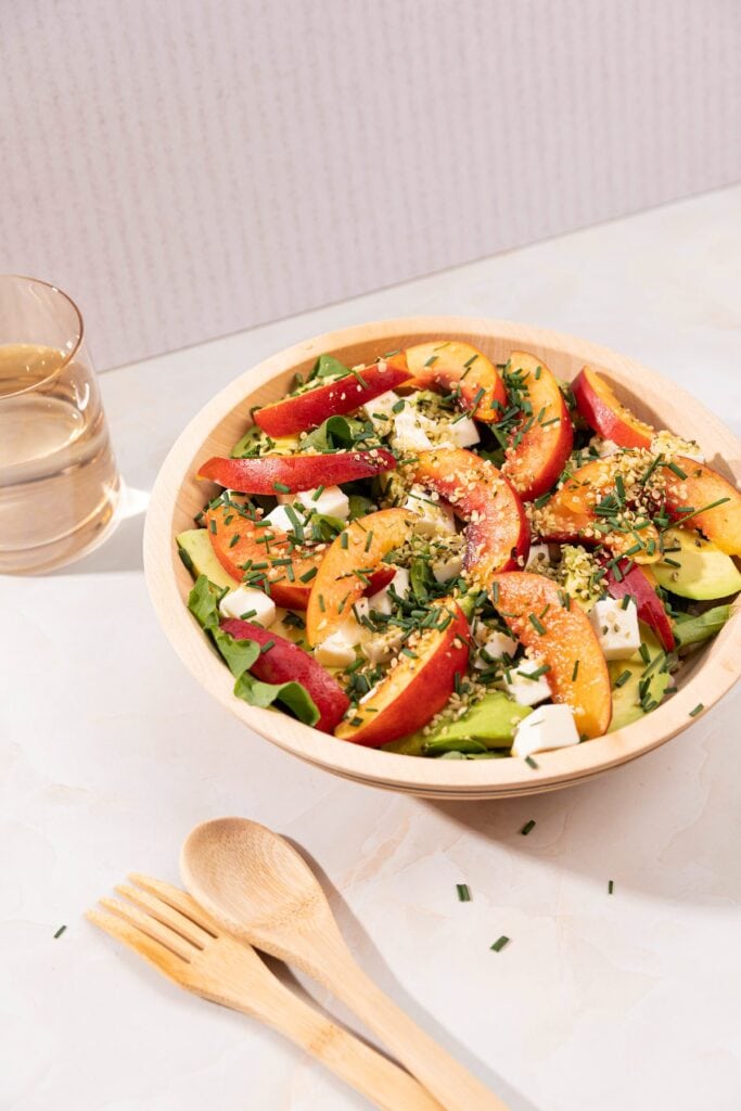The nectarine salad is in the wooden bowl. There are a glass of water and wooden spoon and fork next to it | Hurry The Food Up
