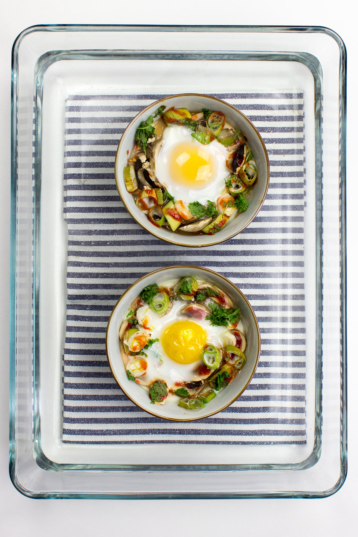 Two bowls of Mushroom Oeuf Cocotte, baked with vegetables and garnished with herbs, rest on a striped cloth inside a glass tray.