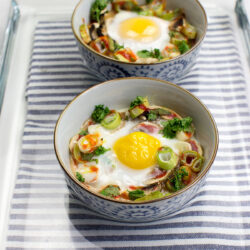Two bowls of Mushroom Oeuf Cocotte, baked with vegetables and garnished with herbs, rest on a striped cloth inside a glass tray.