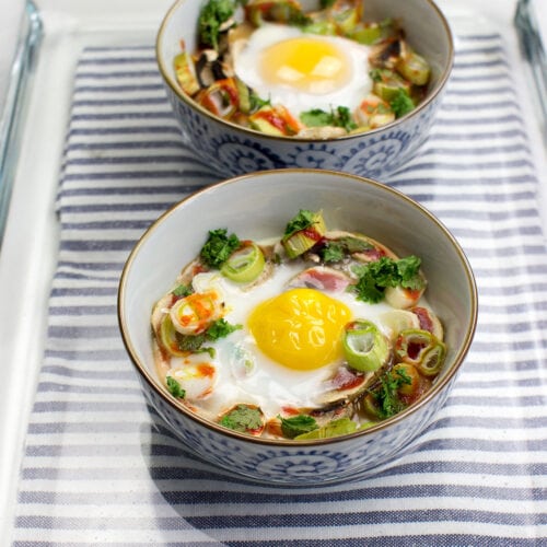 Two bowls of Mushroom Oeuf Cocotte, baked with vegetables and garnished with herbs, rest on a striped cloth inside a glass tray.