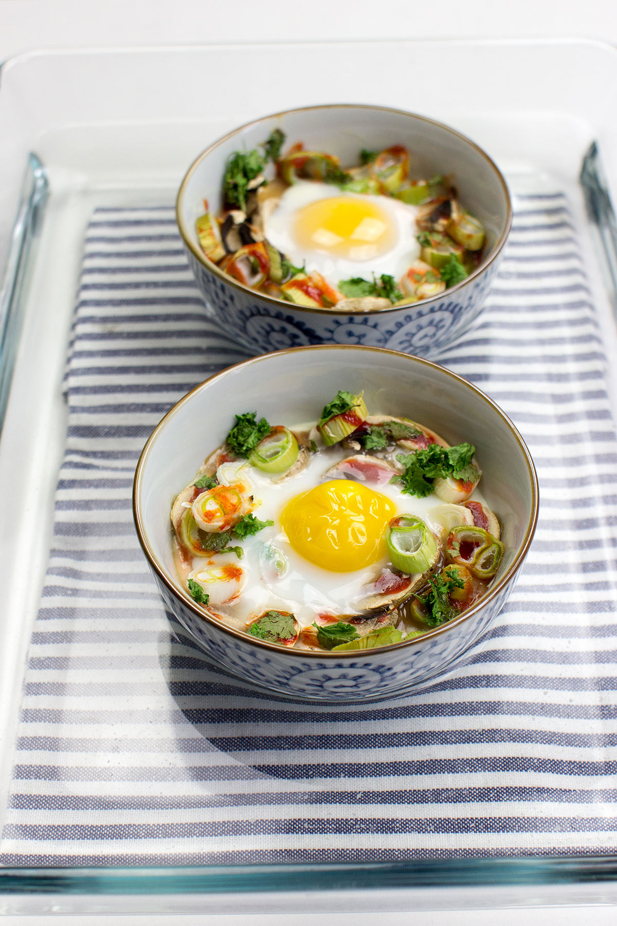 Two bowls of Mushroom Oeuf Cocotte, baked with vegetables and garnished with herbs, rest on a striped cloth inside a glass tray.
