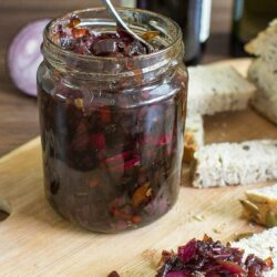 A jar of red onion chutney with a spoon in it, sat on a wooden chopping board. Next to the board are some slices of bread | Hurry The Food Up