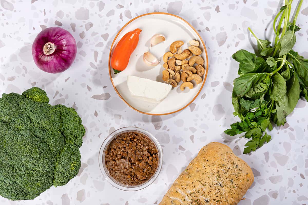 The Oven Roasted Broccoli Halloumi Traybake ingredients like broccoli, red onion, cashews, basil, lime, lentils, bread etc. are on a grey surface | Hurry The Food Up