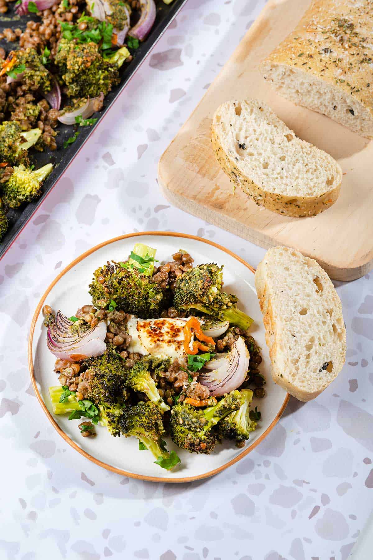 The Oven Roasted Broccoli Halloumi is served on a white plate next to the baking tray and wooden board with bread slices | Hurry The Food Up
