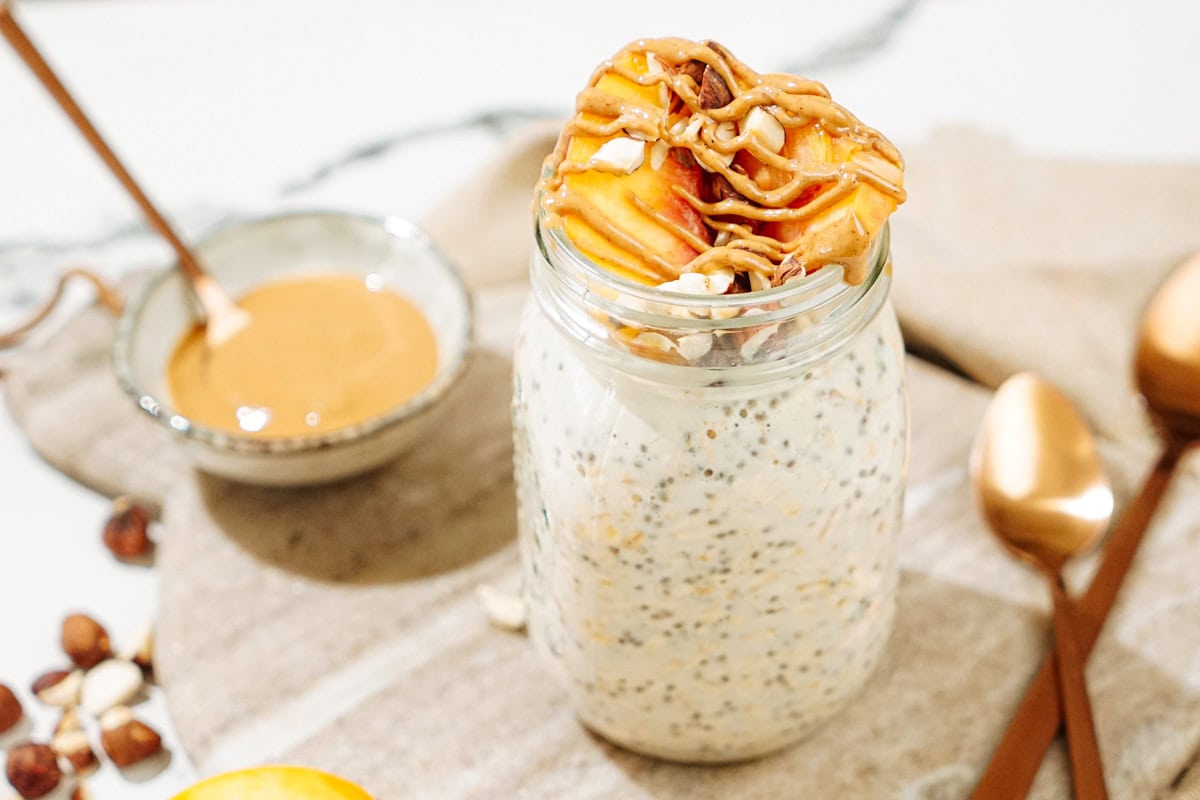 the Mason jar with overnight oats is on the marble cut board. There are some hazelnuts, two bronze spoons, a tiny bowl with peanut butter and a small dish with some slices of peach | Hurry The Food Up